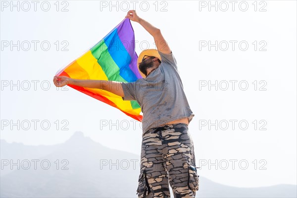 A gay person with a gray t-shirt and white hat waving the LGBT flag in a cloudy sky