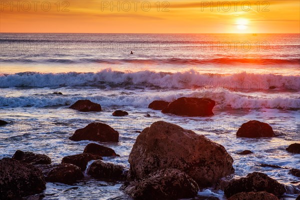 Atlantic ocean sunset with waves and rocks and surfers silhouettes in water at Costa da Caparica