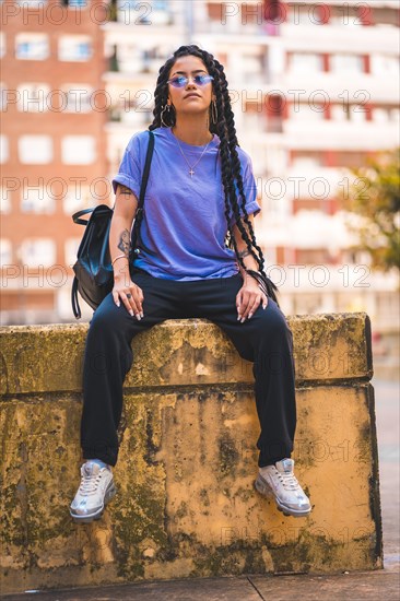 Dark-skinned young woman with long braids and purple glasses sitting on a bench in the city