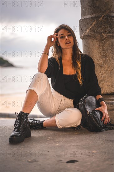 Lifestyle in the city with a blonde girl in white pants and a leather jacket near the beach. Sitting on the shallows of a beach at sunset