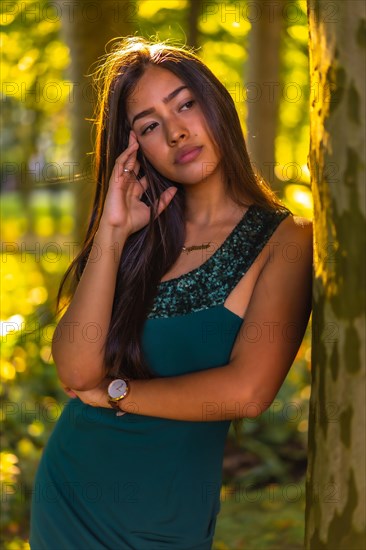 A young brunette Latina with long straight hair leaning against a tree in a green dress. Portrait with a sweet and sensual look