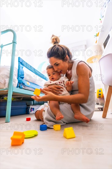 Young Caucasian mother playing with her in the room with toys. Baby less than a year learning the first lessons of her mother. Mother playing with her son and hugging him lovingly