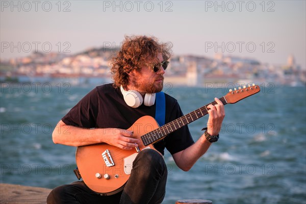 Hipster street musician in black playing electric guitar in the street on sunset on embankment with Lisbon in background. Portugal