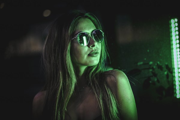 Caucasian brunette girl with sunglasses looking at a light green leds. Night urban photography