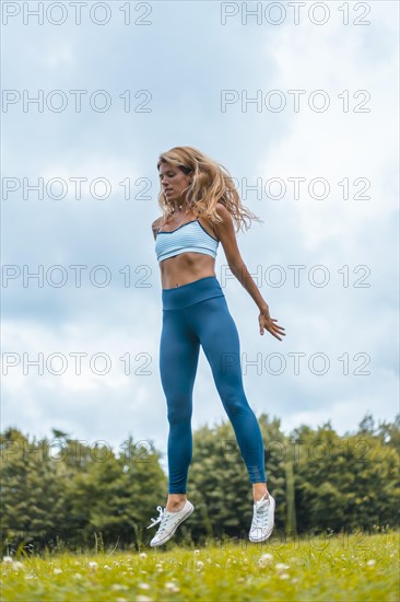 Fitness session with a young blonde caucasian woman exercising in nature with a blue maya on her feet and a white short shirt. Squatting and jumping