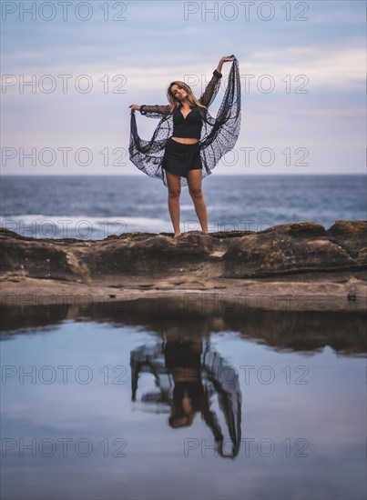 Summer lifestyle with a young brunette Caucasian woman in a long black transparent dress on some rocks near the sea on a summer afternoon. Lifting the dress and smiling