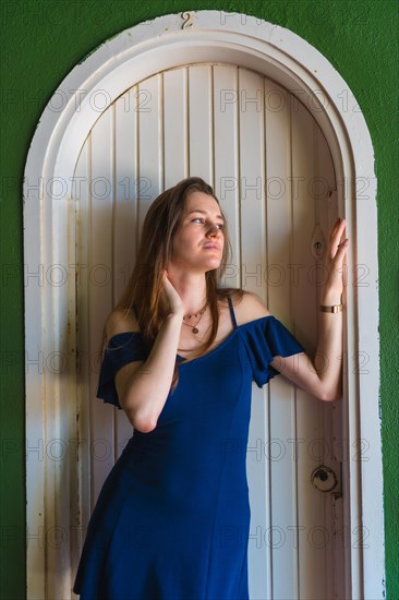 A young redhead Caucasian girl smiling in a blue dress next to a white door of a green house