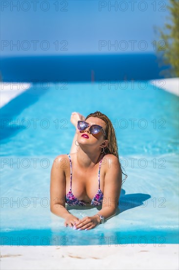 Summer lifestyle in an infinity pool with a young blonde Caucasian woman in a pink and purple bikini with sunglasses. Lying in the pool water with the ocean in the background