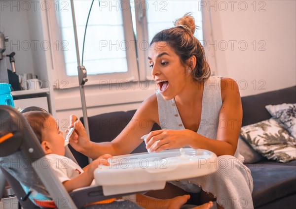 Young Caucasian mother feeding her son a yogurt while sitting in the highchair. Teleworking and caring for your child