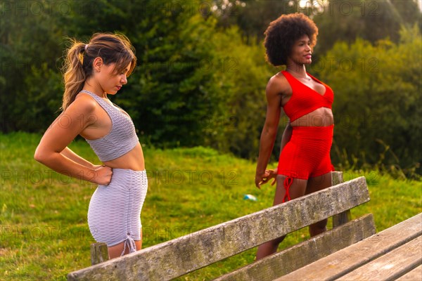 Caucasian blonde girl and dark-skinned girl with afro hair doing stretching before starting sports in the park. Healthy life