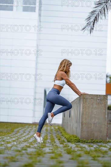 Fitness and yoga session with a young blonde Caucasian instructor dressed in a casual outfit with blue Maya and a white T-shirt. Exercising with one leg roblandola on a white wall background