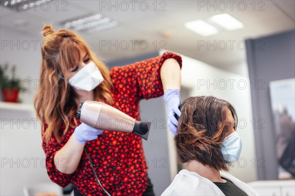 Blonde hairdresser with mask and gloves drying the client's brown hair with a hairdryer. Reopening with security measures for hairdressers in the Covid-19 pandemic