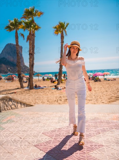 A Caucasian redhead dressed in white and with a straw hat walking along the beach of Calpe in summer