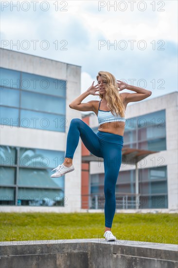 Fitness session with a young blonde Caucasian woman exercising in a park next to offices with a blue maya on her feet and a white short shirt. Performing leg exercises