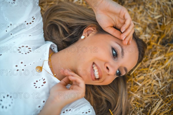 A young blonde Caucasian woman in a white dress in a field of dry straw atop a haystack