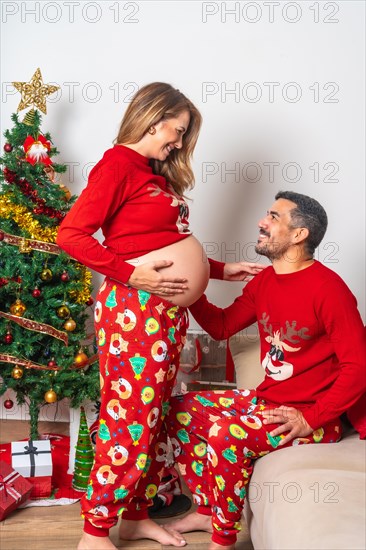 Young couple with decorations and red Christmas clothes looking at each other and very happy for the baby boy who is coming to the family. Family with pregnant woman