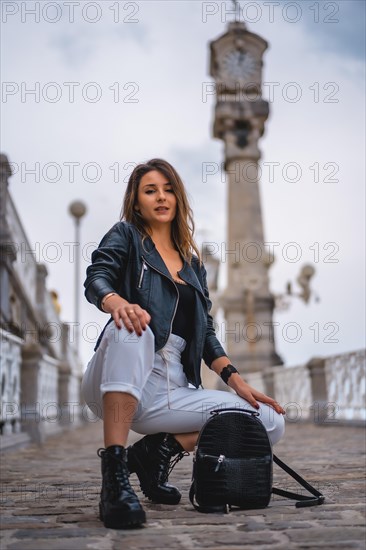 Lifestyle in the city with a blonde girl in white pants and a leather jacket. Crouched on a slope with her bag