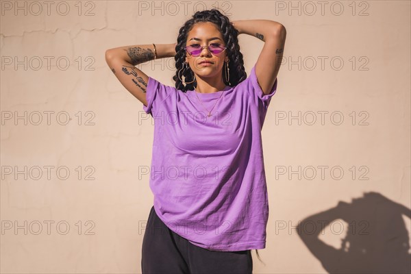 Urban session. portrait of young dark-skinned woman with long braids wearing purple glasses on a plain cream-colored background