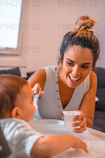 Young Caucasian mother feeding her son a yogurt while sitting in the highchair. Teleworking and caring for your child