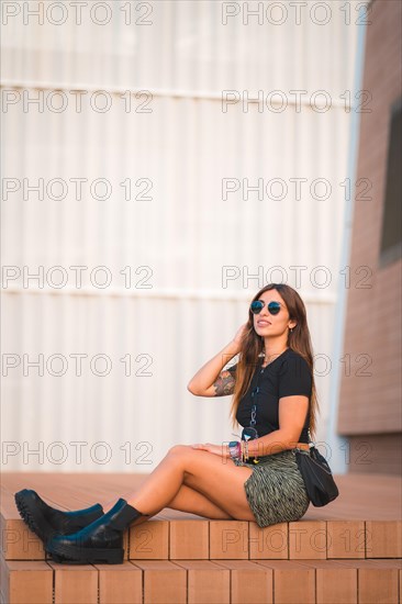 Street style of a young caucasian brunette sitting in the city enjoying verana with a white wall background