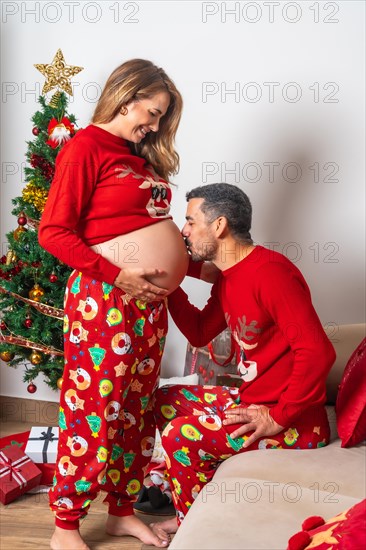 Young couple with decoration and red Christmas clothes kissing pregnant girlfriend's belly. Family with pregnant woman