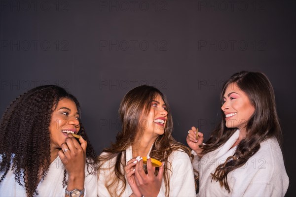 Studio photo with grey background of multi-ethnic women in bathrobe having fun eating fruit