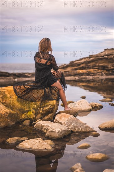 Summer lifestyle with a young brunette Caucasian woman in a long black transparent dress on some rocks near the sea on a summer afternoon. Sitting on top of a rock in a natural pool