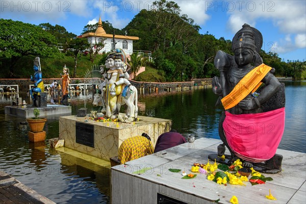 Right Hindu deity statue figure sculpture of goddess deity of Lord Muruga left beside Dattatreya triune figure Brahma Vishnu beside Lord Shiva in background woman Shakti of Shiva Hindu mother goddess mother of Ganesha in lake sacred Hindu lake Ganga Talao at religious site largest Hindu sanctuary sanctuary for religion Hinduism outside India for devout Hindu Hindus