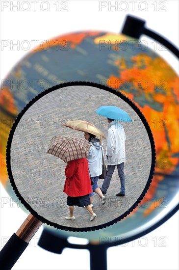 Tourists walking in the rain with umbrellas in summer seen through magnifying glass held against illuminated terrestrial globe