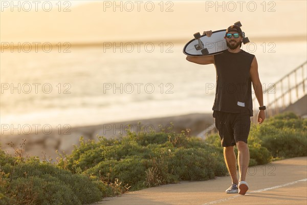 A man walking on the road near by beach holding a skateboard. Mid shot
