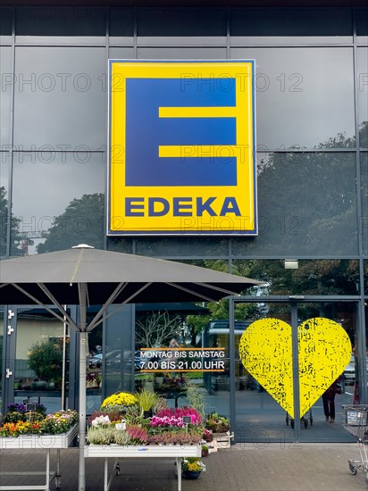 Logo with letter and lettering above entrance to supermarket below note on opening hours Monday to Saturday 7.00 a.m. to 9.00 p.m. to the right stylised heart of retail chain supermarket chain Edeka formerly during German colonial times Einkaufsgenossenschaft der Kolonialwarenhaendler