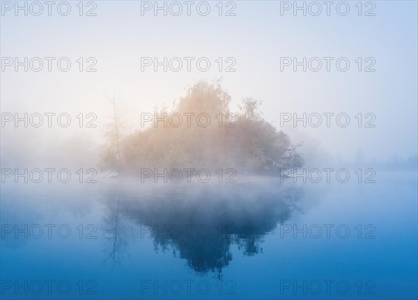 Foggy sunrise at a lake. Island and trees in blue and orange light