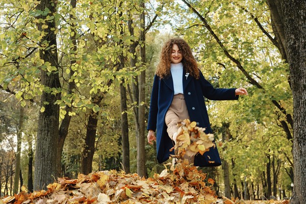 Low angle view of young happy woman kicking up autumn leaves in park and laughing