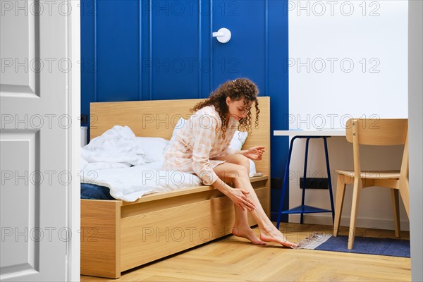 Young woman in bathrobe sitting on bed and applying moisturising cream on her legs