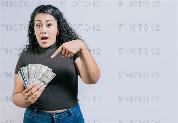 Amazed young woman holding and pointing dollars banknotes isolated. Surprised latin girl holding dollar cash isolated