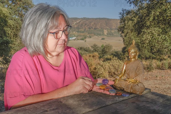 Older white-haired woman with chakra stones in her hands next to a Buddha statue