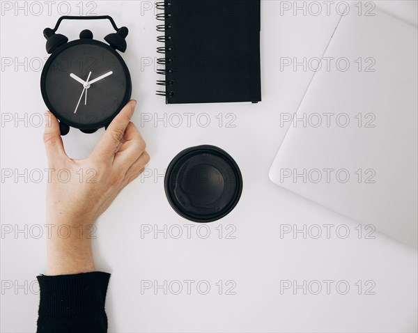 Close up person s hand holding black alarm clock spiral notepad laptop takeaway coffee cup white desk