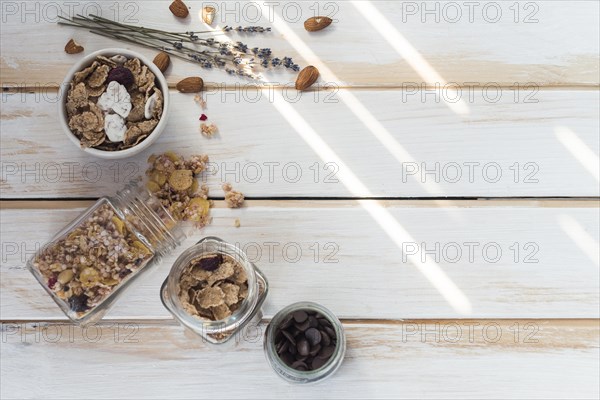 Jar spilled granola near cornflakes dry fruits chocolate chips wooden plank