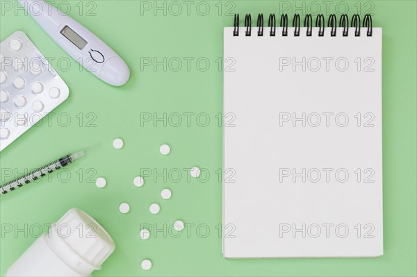 Pills thermometer syringe plastic bottle near spiral notepad green background