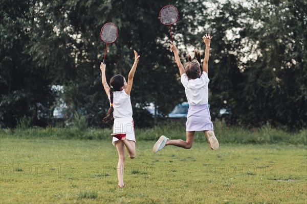 Rear view two girls jumping park holding badminton