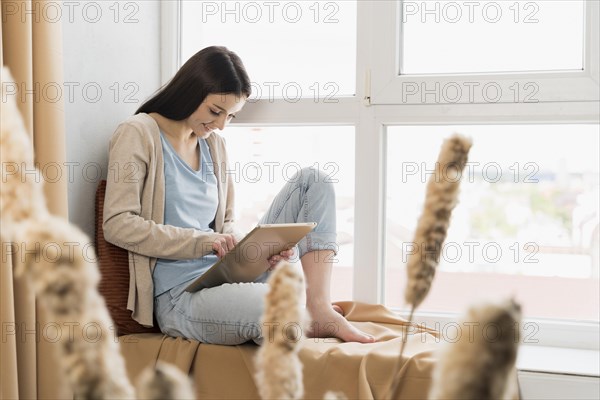Side view woman sitting window working tablet
