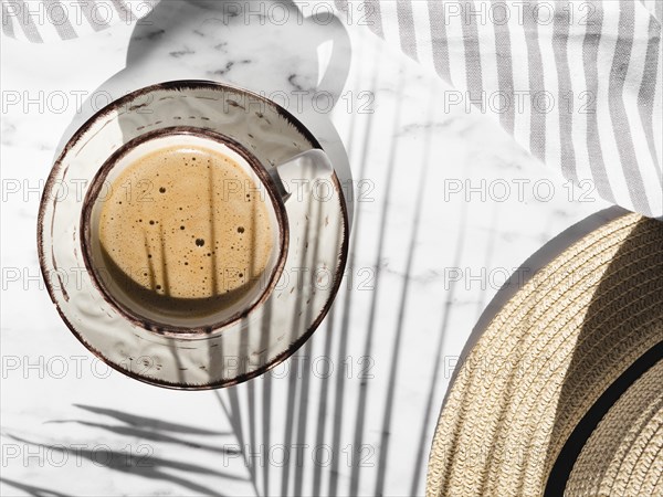 White cup with red shapes filled with creamy coffee white background with striped grey white cloth covered by ficus leaf shadow hat