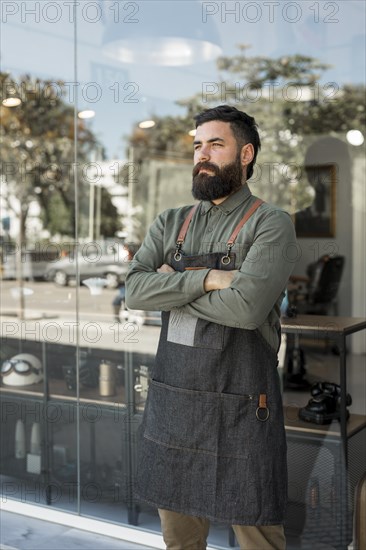 Bearded serious hairdresser standing outside near barbershop