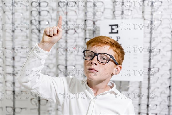 Boy with spectacle pointing upward direction standing against eyeglasses display background