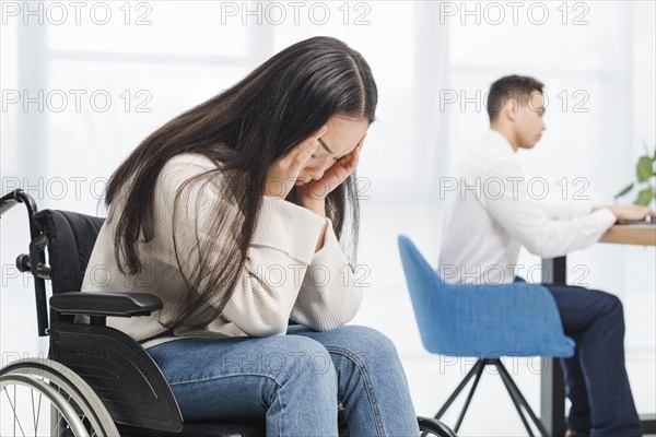 Frustrated young woman suffering from headache sitting wheel chair front businessman working office