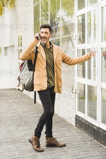 Modern smiling man with his backpack talking mobile phone while opening glass door