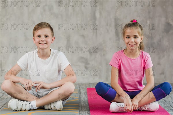 Portrait smiling girl boy sitting exercise mat with their crossed legs front wall