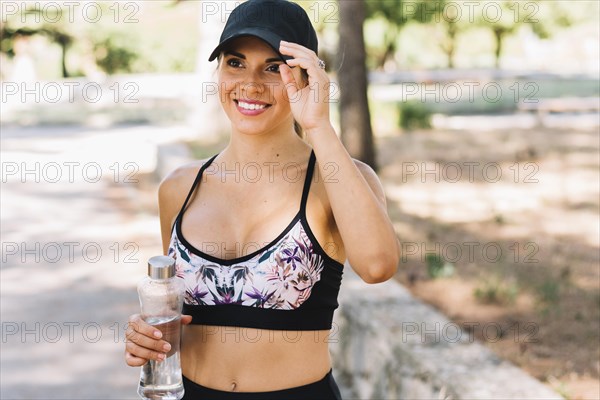 Portrait smiling sporty young woman wearing black cap holding plastic water bottle