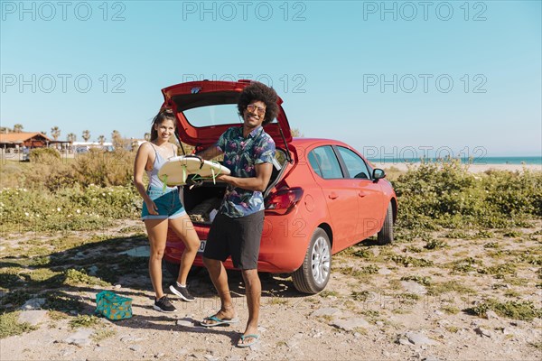 Young black man holding surfboard near car by seashore