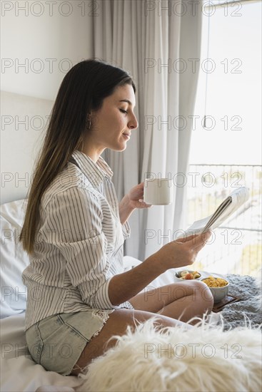 Young woman sitting bed reading newspaper while drinking coffee